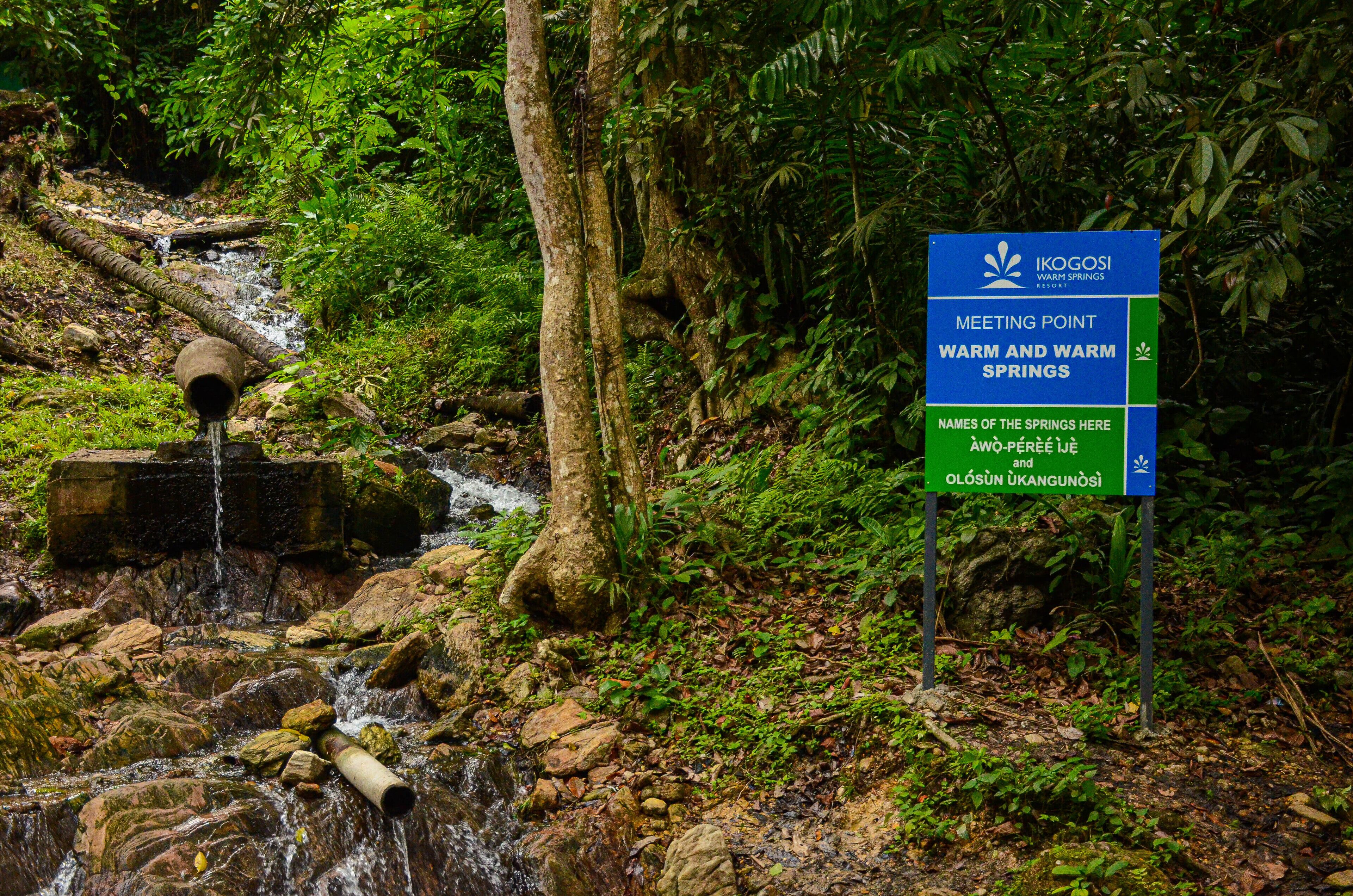 A sign for Ikogosi Warm Springs Resort with a natural waterfall and lush greenery.
