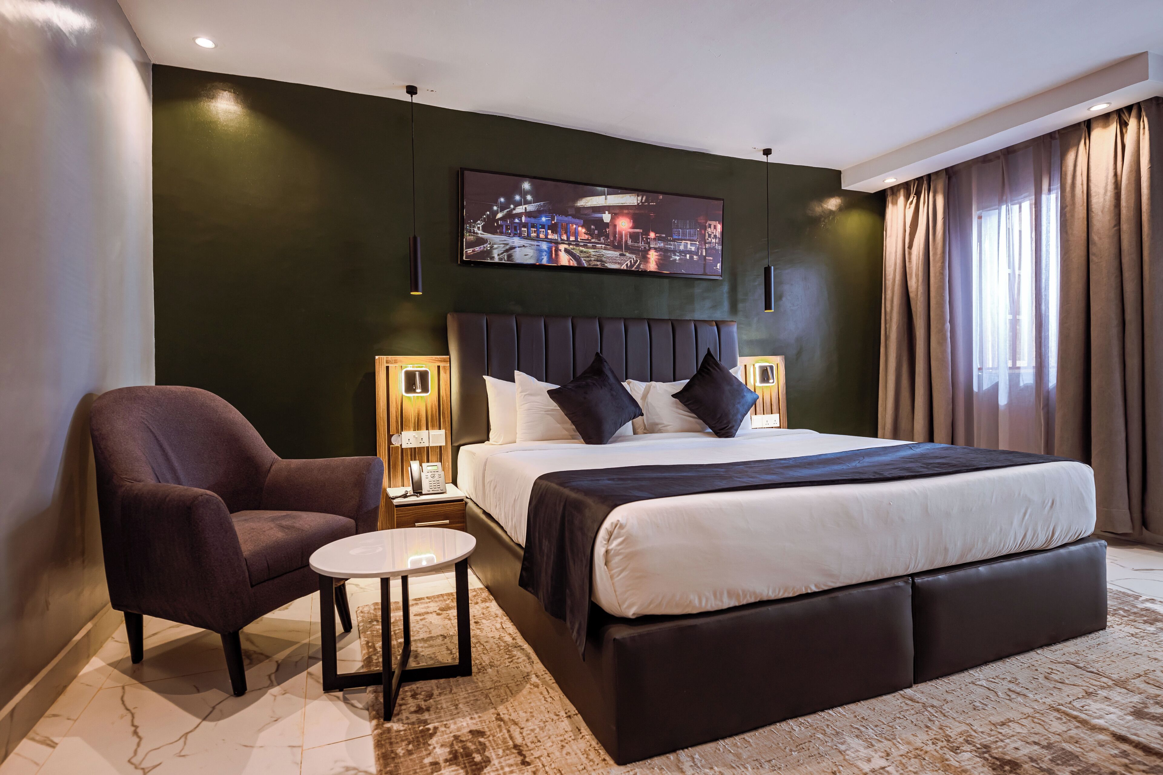 A modern hotel room with a large bed, a chair, a small table, and a nightstand with a lamp.
