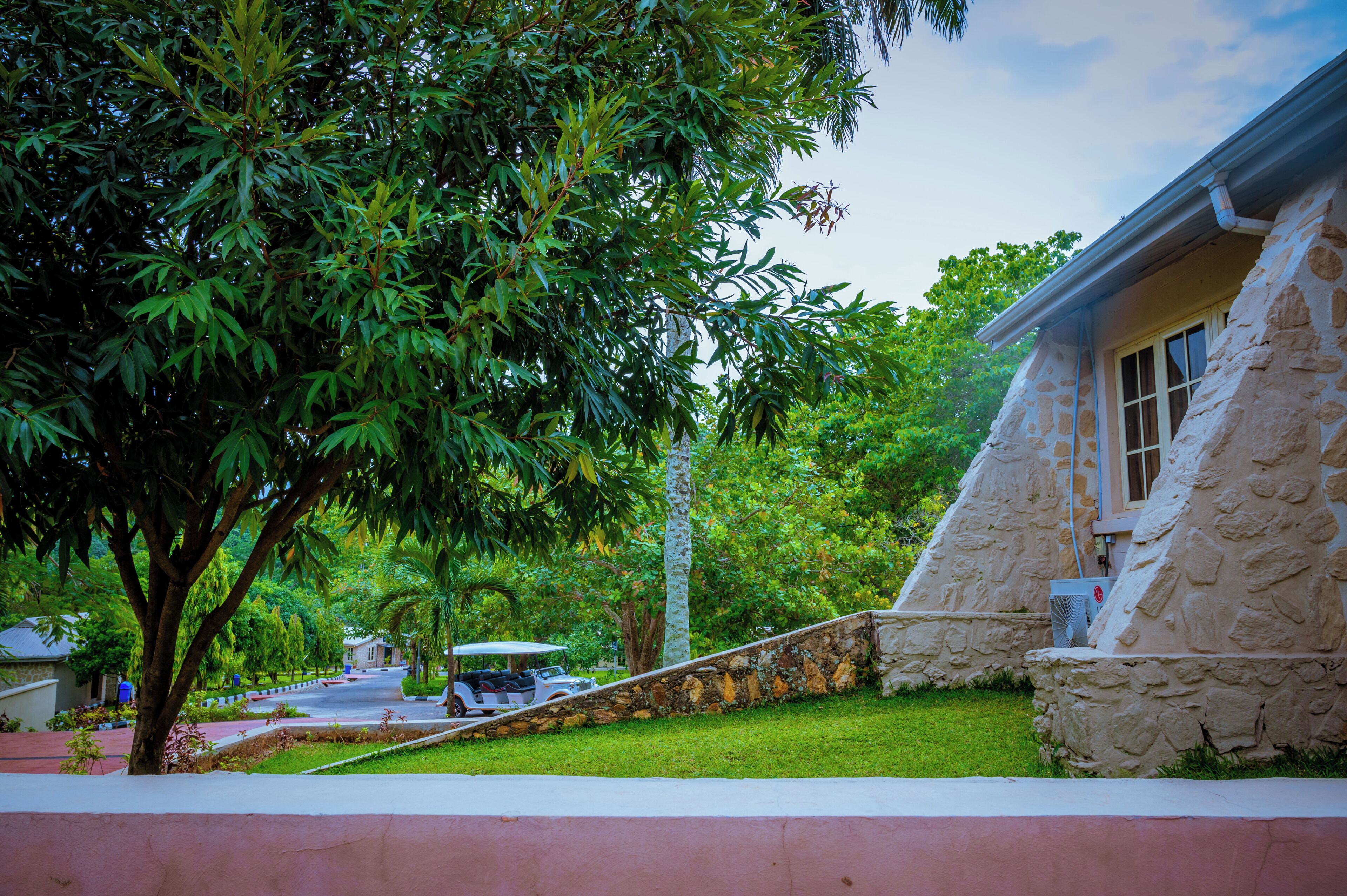 A stone building with a sloped roof, surrounded by lush greenery and a paved driveway.