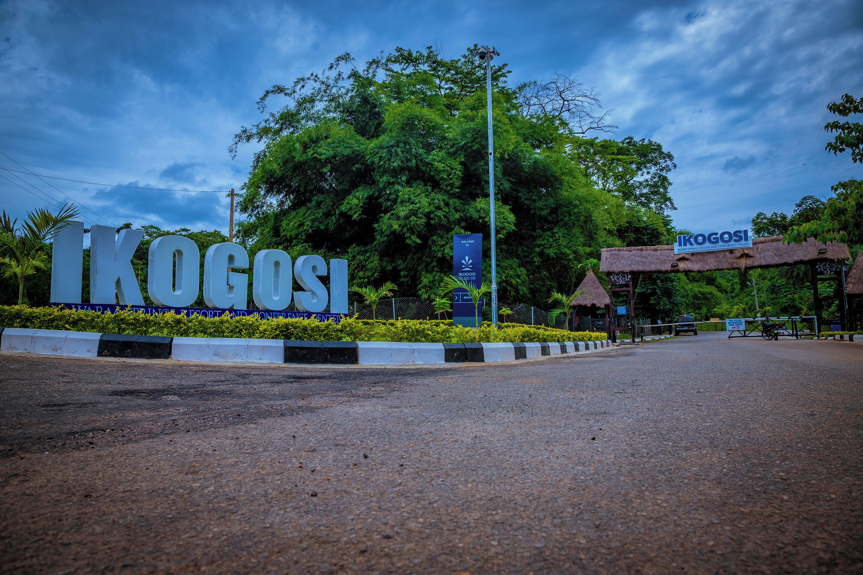 A road leading to a sign with "IKOGOSI" written on it, surrounded by greenery and a clear sky.
