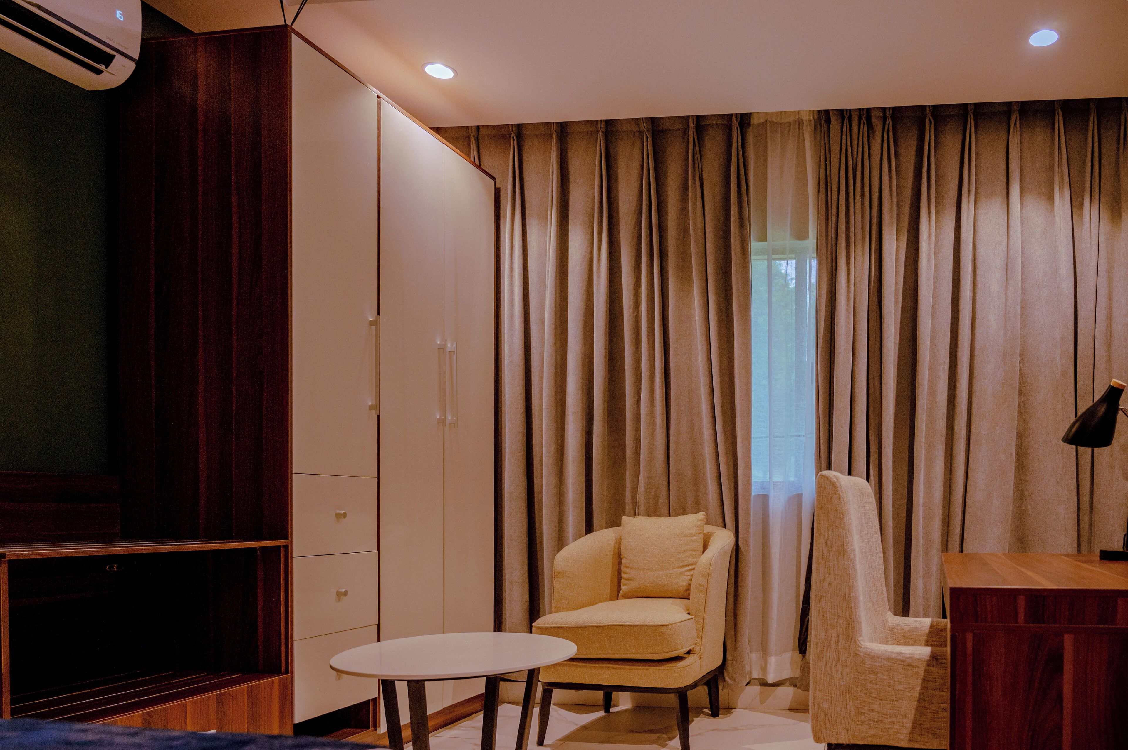 A hotel room with a large wardrobe, a small round table, two armchairs, and a desk with a lamp.