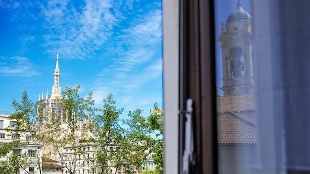Une vue d’un bâtiment historique, doté d’une haute flèche et d’une tour horloge, dont le reflet se mire dans une fenêtre.