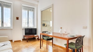 Une pièce moderne et bien éclairée, avec une table à manger en bois, deux chaises, un canapé, un téléviseur et un coin cuisine visible à travers une porte coulissante.