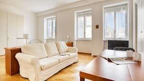 Une pièce moderne et bien éclairée, dotée d’un plancher en bois, d’un canapé beige, d’une table en bois et de grandes fenêtres ornées de rideaux légers.