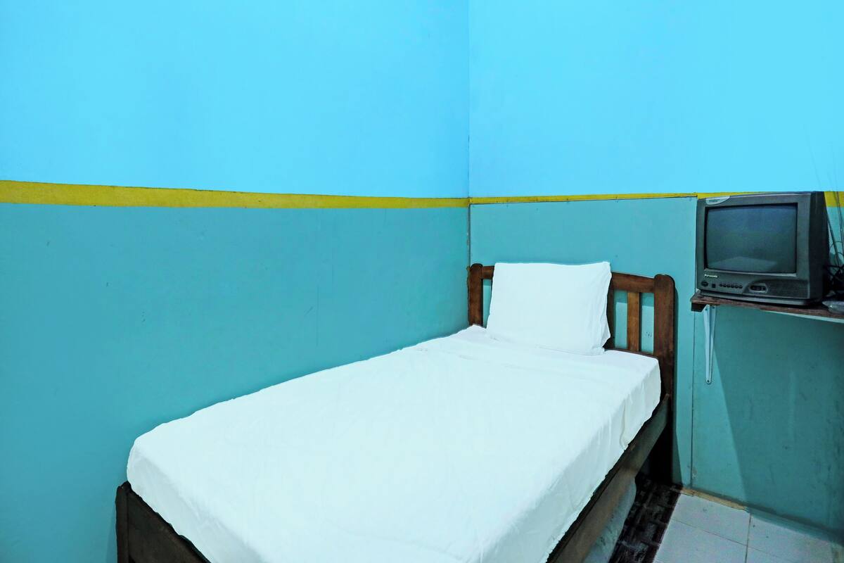A small, single-bed room with a wooden headboard and a white mattress. A television is mounted on the wall to the right of the bed.
