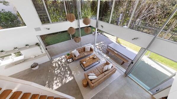 A modern interior with a large glass door leading to a garden, a sofa set, and hanging pendant lights. - 25 Whitehaven Parade (Yaroomba)