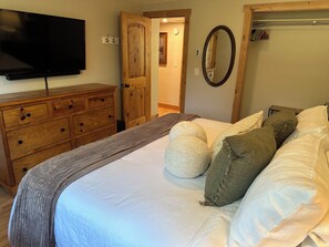 Un dormitorio con cama, cómoda, televisor, espejo y puerta que da a otra habitación.