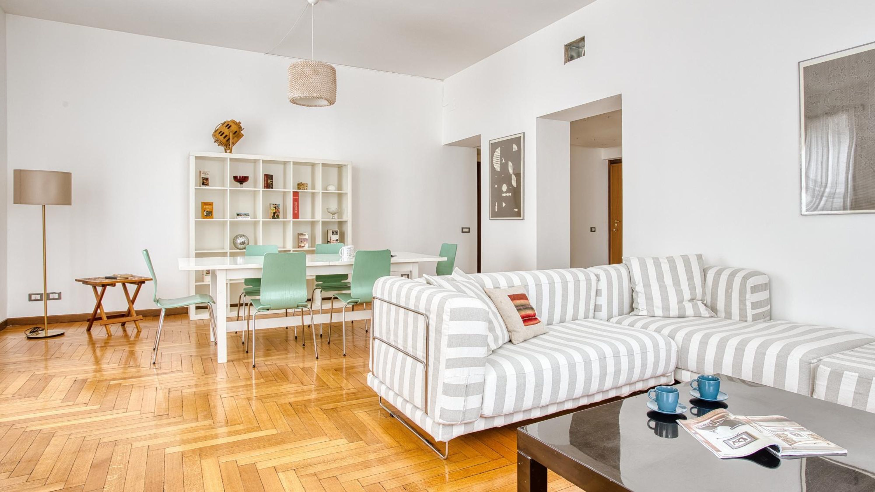 A modern living room with a striped sofa, a dining area with a white table and green chairs, and a bookshelf with various items.