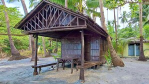 A wooden structure with a triangular roof, a bench, and a door, surrounded by palm trees and a sandy ground. - Playa La Caleta Bataan (Morong)