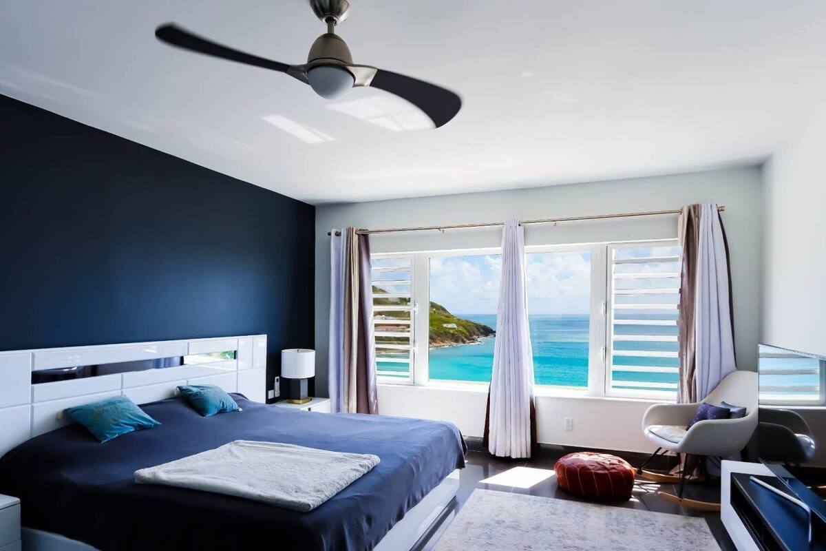 A modern bedroom with a large bed, a ceiling fan, a desk with a chair, and a view of the ocean through the window.