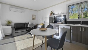 Une cuisine moderne équipée d’un lave-vaisselle, un coin repas avec une table ronde et des chaises, un canapé en cuir noir et une fenêtre donnant sur la verdure.