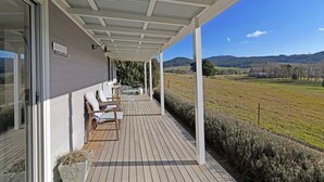 Un espace extérieur abrité, doté d’une terrasse en bois, de chaises blanches et offrant une vue sur un paysage rural.