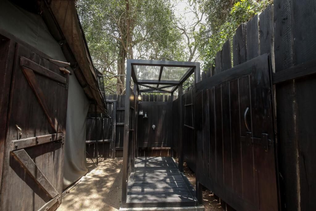 A narrow pathway leading to a covered area with a metal frame and glass roof, surrounded by wooden fences and trees.