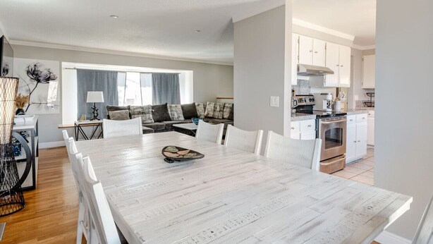 Een ruime eethoek met een lange tafel en witte stoelen, een moderne keuken met witte kasten, en een woonkamer met een bank en kussens.