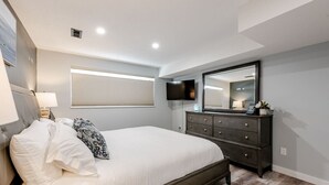 Een slaapkamer met een bed, een dressoir, een televisie en een raam met jaloezieën.