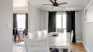 Een eetkamer met een witte tafel en stoelen, een plafondventilator en een uitzicht op buiten door grote ramen.