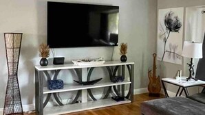 Een woonkamer met een flatscreen televisie, een console tafel, een grijze bank, een kamerkleed met patroon en een gitaar.