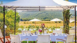 An outdoor dining area with tables set for four, surrounded by white chairs, under a canopy with a view of a green landscape and hills.