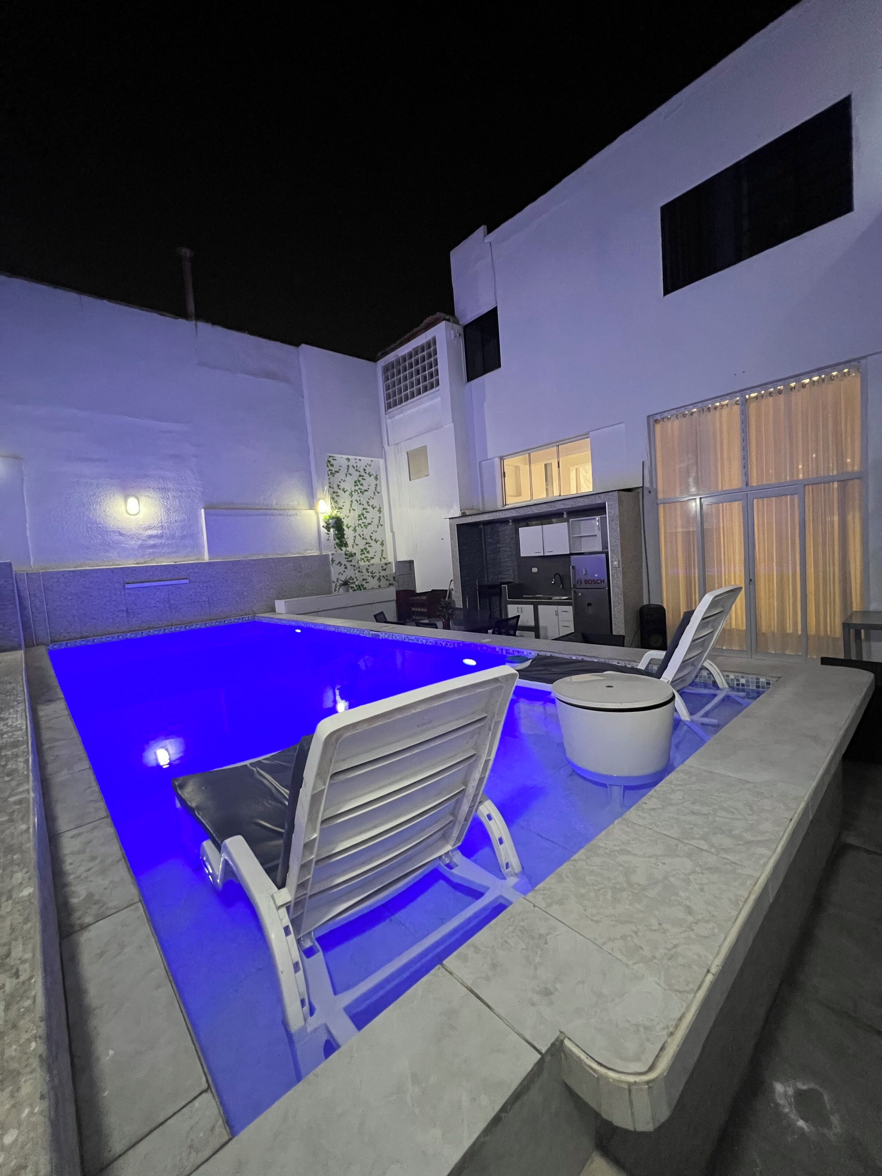 A swimming pool with a blue-lit edge, a white chair, and a white table with a white cover.