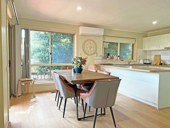 A modern kitchen with a dining area, large windows, and a view of greenery.