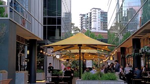 An outdoor dining area with tables and chairs under a large canopy, surrounded by modern buildings. - Ringwood 2BR Home  (Ringwood)