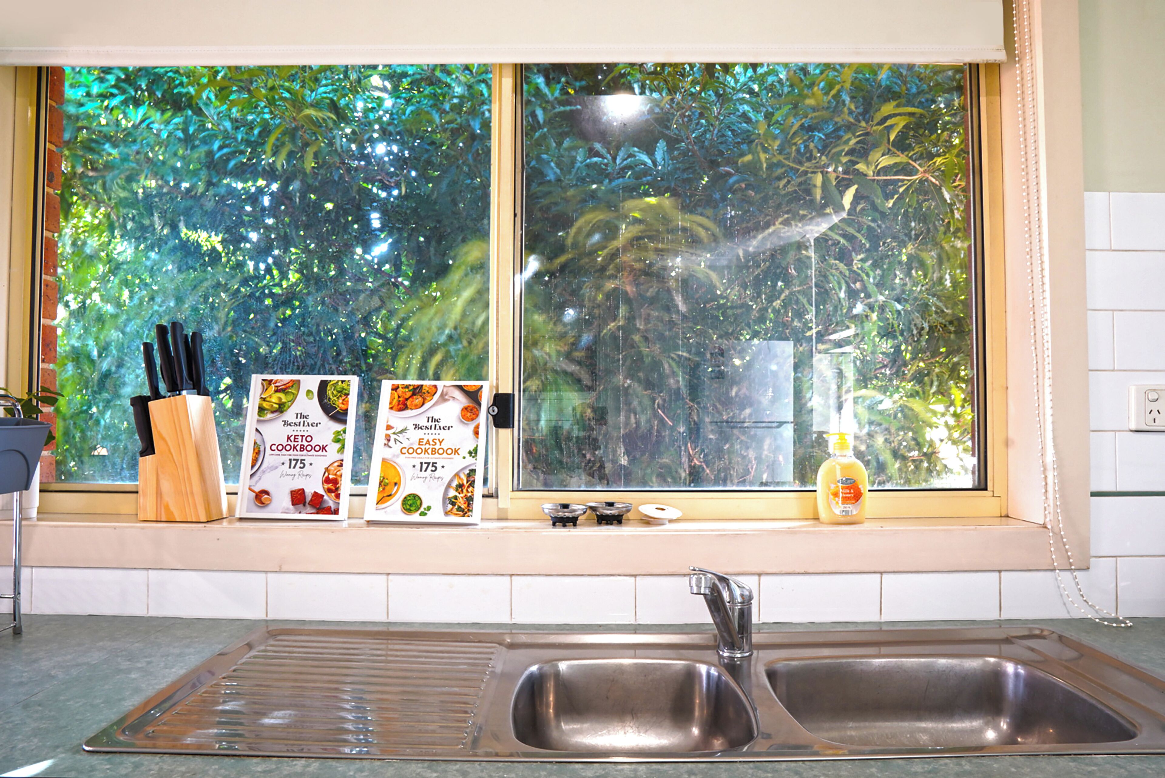 A kitchen with a double sink, a window with a view of greenery, and two cookbooks on the windowsill.