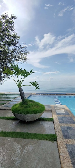 Un bac à fleurs en béton, avec un arbre et de l’herbe, placé à côté d’une piscine à motifs à carreaux, sous un ciel dégagé.