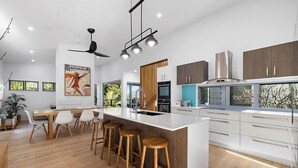 A modern kitchen with a central island, bar stools, and a dining area with a white table and chairs. - Stunning House in peaceful coastal setting (Coolum Beach)