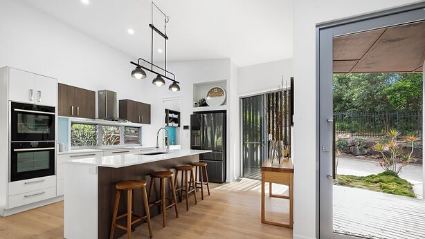 A modern kitchen with a central island, bar stools, a refrigerator, and a large window overlooking a garden. - Stunning House in peaceful coastal setting (Coolum Beach)