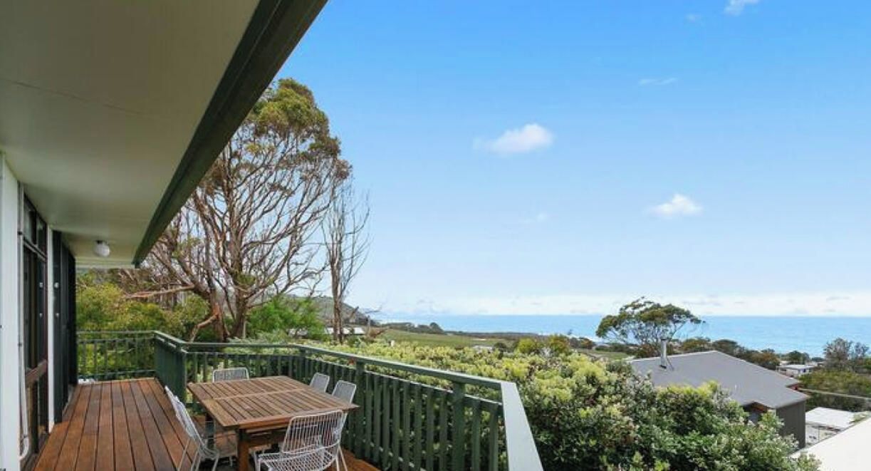 A balcony with a table and chairs, overlooking a coastal landscape with trees and a view of the sea.