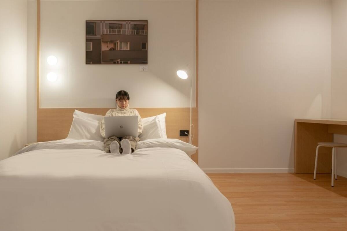 Eine Person sitzt auf einem Bett, zusammen mit einem Laptop, einem Schreibtisch und einem Stuhl in einem Zimmer mit Holzboden und weißen Wänden.