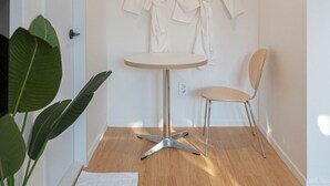 Una stanza con pavimento in legno, un tavolo rotondo con gambe in metallo, due accappatoi bianchi appesi al muro, una sedia e una pianta in vaso.