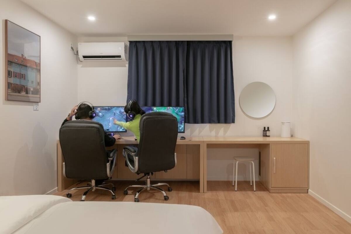 Ein modernes Hotelzimmer mit Schreibtisch aus Holz, zwei Bürostühlen, einem großen Flachbildfernseher, einem runden Spiegel und einem kleinen Hocker.