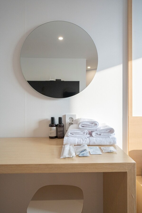 Habitación de hotel con un espejo redondo, una mesa de tocador de madera, toallas blancas ordenadas y artículos de aseo.