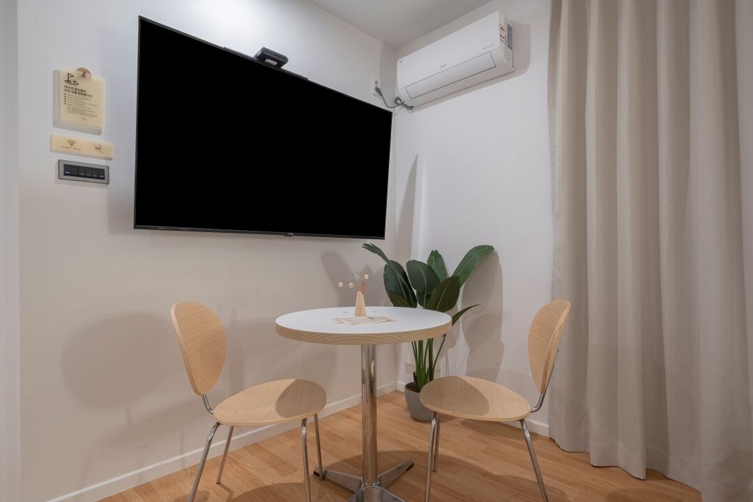 A room with a round table and two chairs, a flat-screen TV mounted on the wall, an air conditioning unit, and a potted plant.