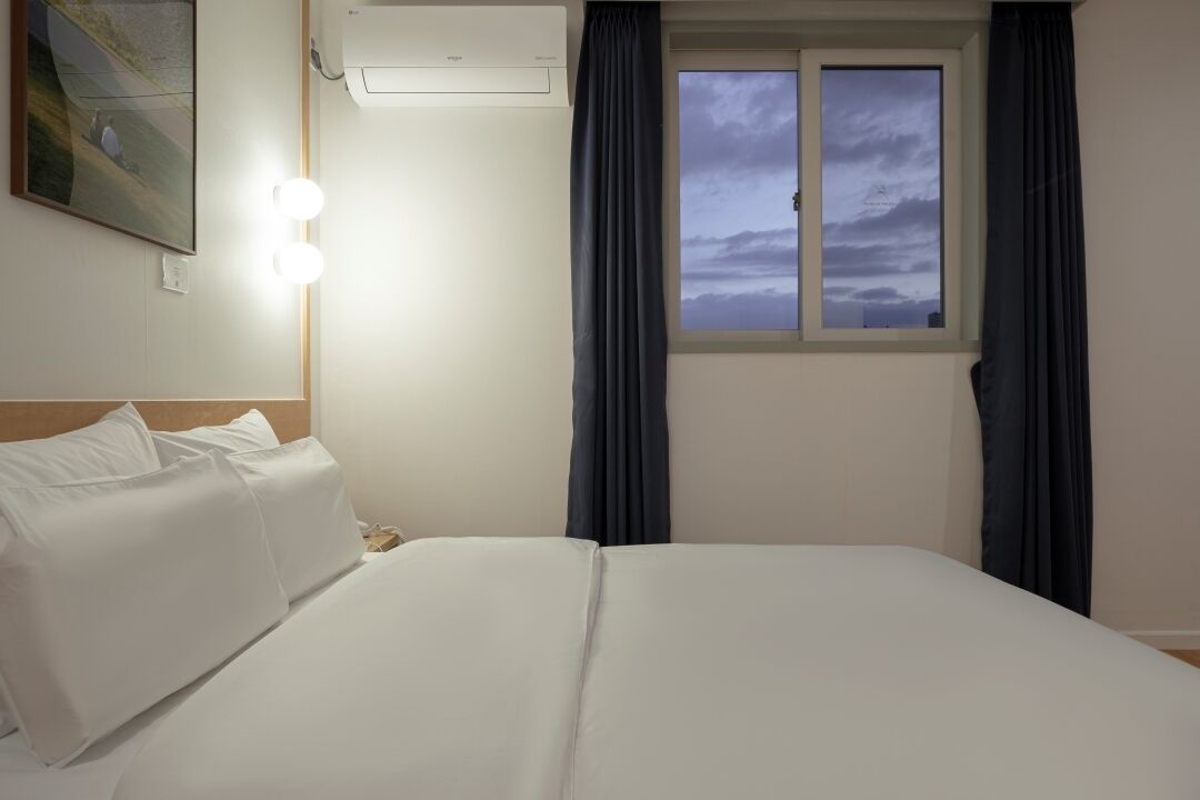 A hotel room with a bed, a painting on the wall, an air conditioning unit, a window with curtains, and a night light.