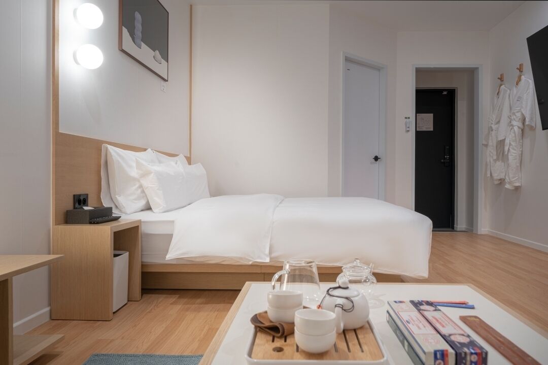 A modern hotel room with a neatly made bed, bedside table with a lamp, and a tray with tea and books.