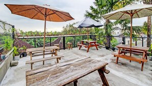 Een terras buiten met houten tafels en banken, twee parasols en potplanten.