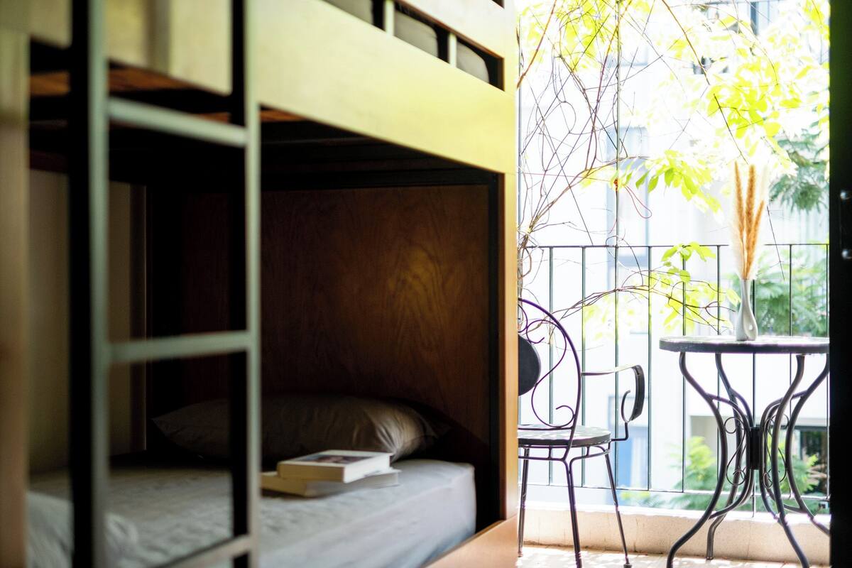 A room with a bunk bed, a small table, and a chair. There is a balcony with a railing and a plant.