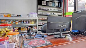 A checkout counter with a computer monitor, keyboard, and scanner. There are shelves with various products in the background.