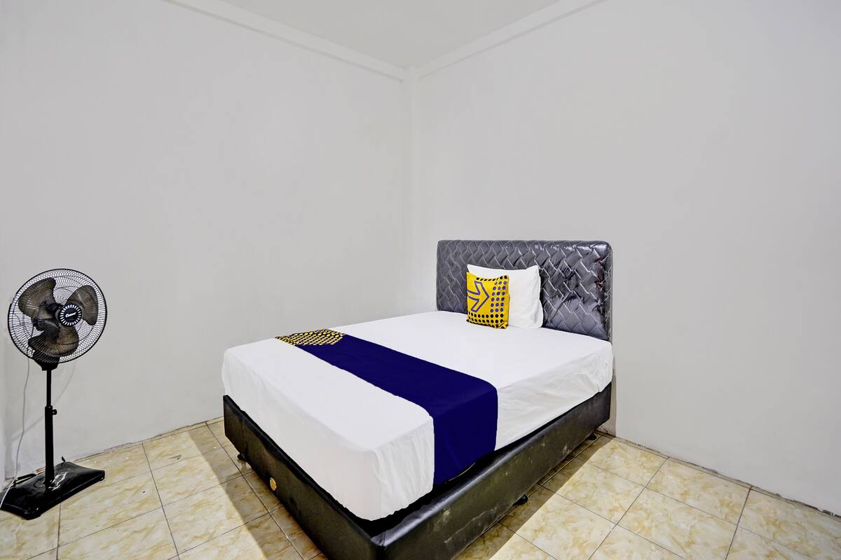 Un lit bien fait, recouvert d’une couverture blanche et bleue, avec un oreiller à motifs géométriques jaunes, et un ventilateur sur pied placé à gauche.