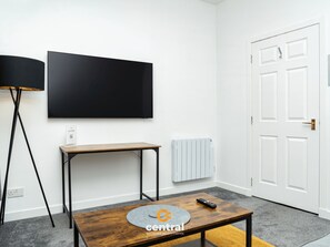 A modern living room with a flat-screen TV mounted on the wall, a small wooden table with a remote control, a standing floor lamp, and a radiator.