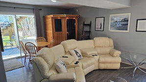 A modern living room with a beige sectional sofa, a glass coffee table, and a wooden cabinet. There are framed pictures on the wall and a view of the outdoors through a glass door.
