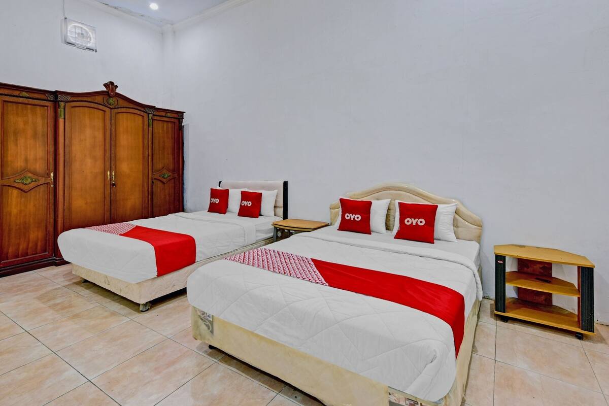 Une chambre d’hôtel avec deux lits, chacun recouvert d’une courtepointe rouge et blanche et agrémenté d’oreillers, ainsi qu’une armoire en bois et une petite table de chevet en bois.