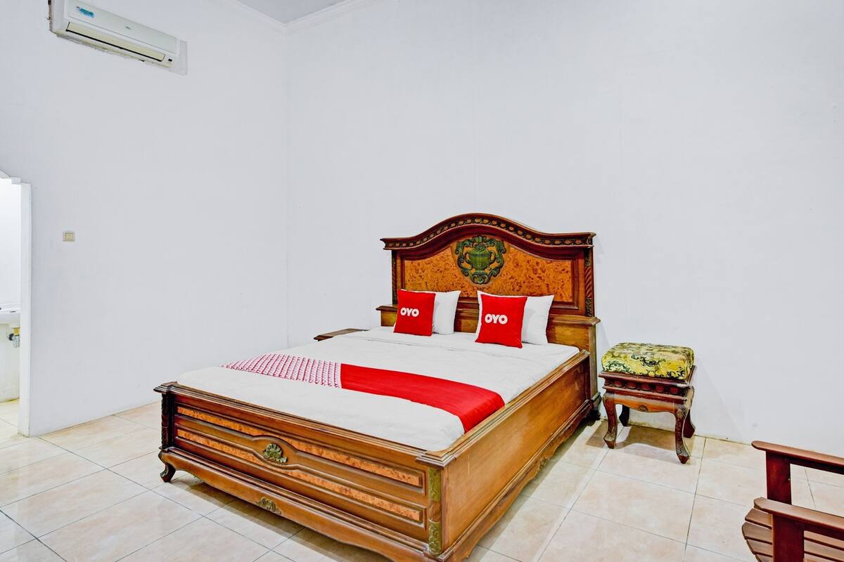 Un lit en bois avec une couvre-lit rouge et blanche et des coussins assortis, une petite table de chevet en bois avec un coussin à motifs, et un mur blanc agrémenté d’une tête de lit décorative.