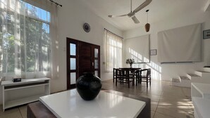 A modern living room with a large window, a ceiling fan, a dining area, and a white coffee table with a black vase.
