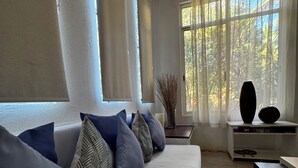 A modern living room with a sofa, decorative pillows, a vase with branches, and a view of trees through large windows.