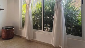 A room with two large windows, white curtains, a wicker basket, and a tiled floor.