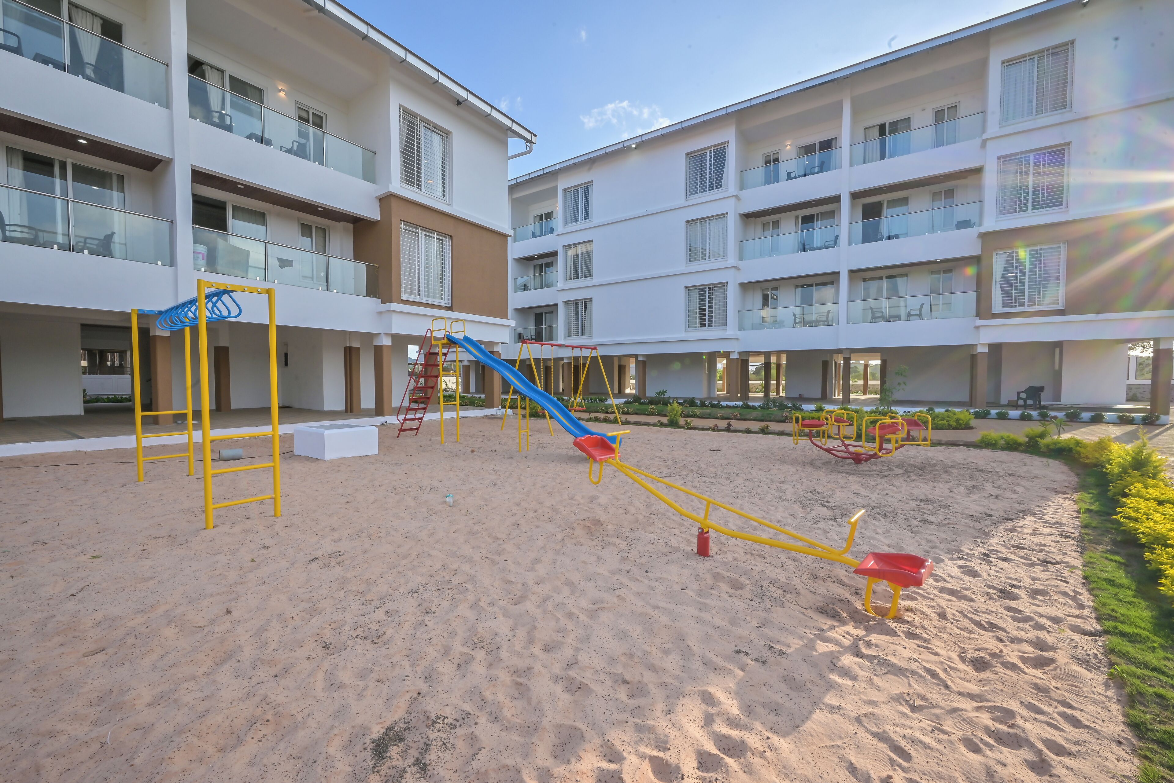A playground with a slide and swings, surrounded by a sandy area and a building with balconies.