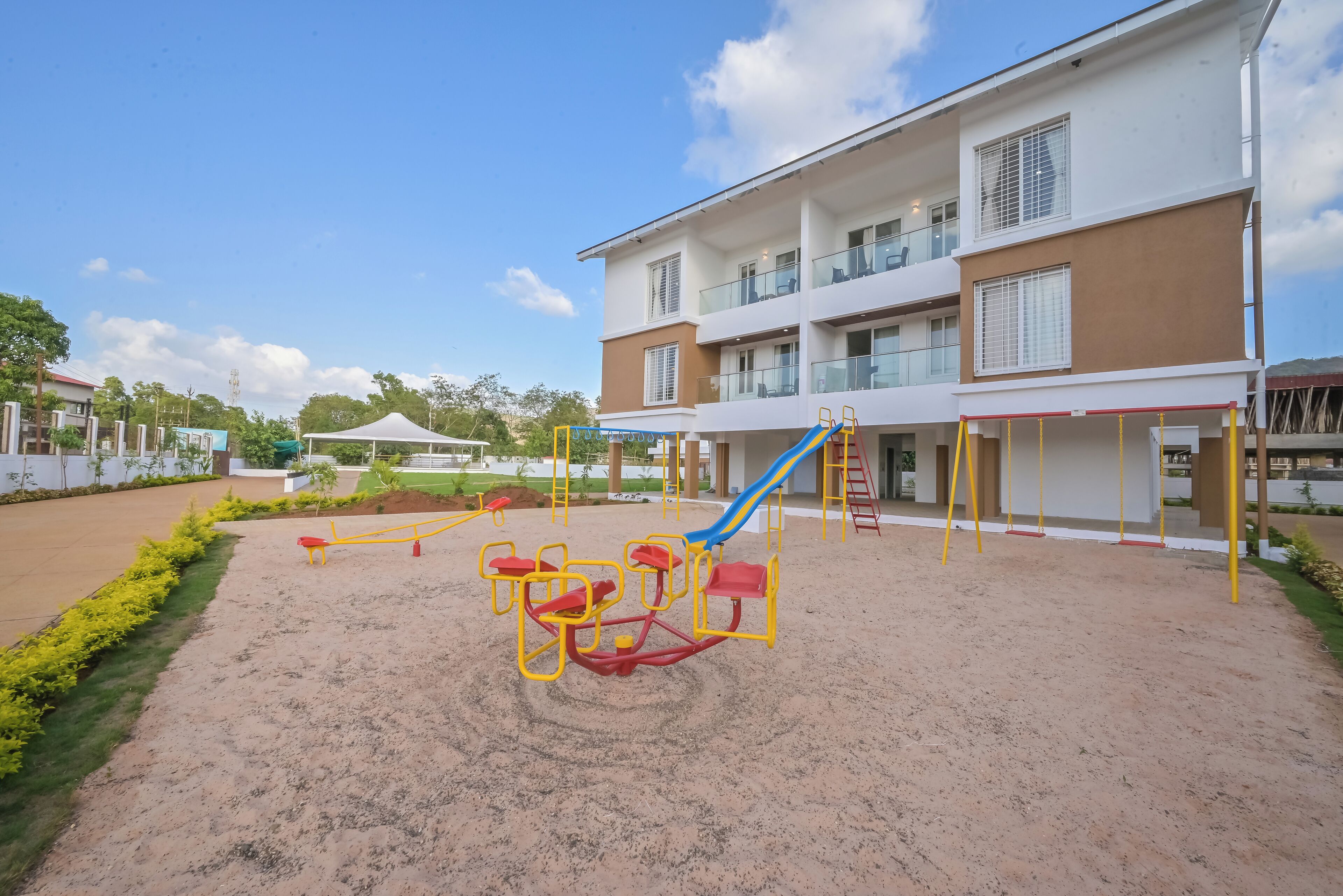 A playground with swings and a slide, a building with multiple floors and balconies, and a clear sky.